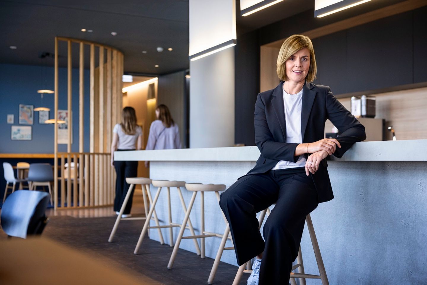 Dr. Eva Griewel, CFO of the EOS Group, is sitting in a social space on a high stool, wearing a black blazer.