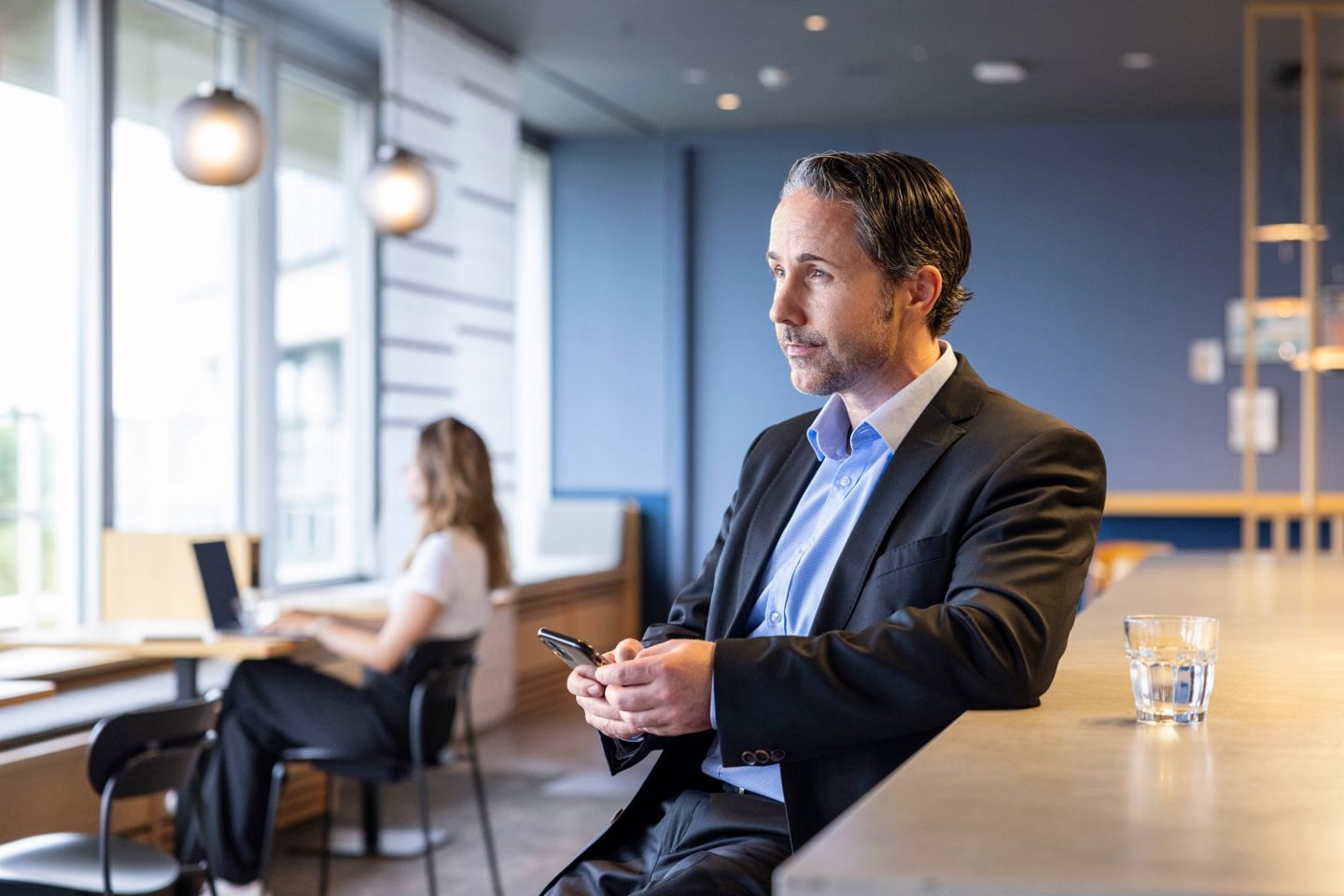 Marwin Ramcke is standing at the counter in a modern social space, wearing a black jacket, holding a glass of water and his phone.